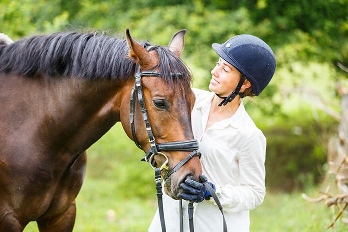 Woman wearing riding helmet brushing brown horse and smiling